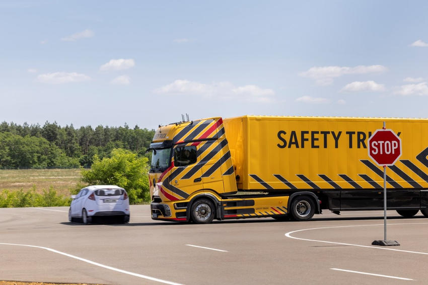Gelber Mercedes-Benz Safety Truck nähert sich auf einem Testgelände einer Kreuzung, während ein weißer Pkw vorbeifährt.