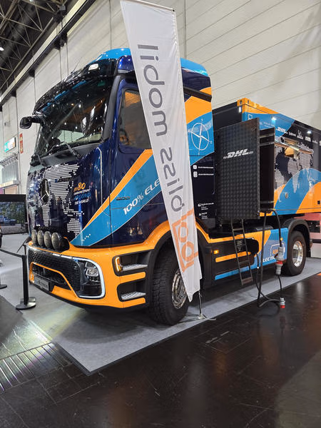 Blau-orangefarbener Mercedes-Benz eActros 600 mit Expeditionsaufbau und Ladeeinrichtung hinter dem Fahrerhaus in einer Messehalle.