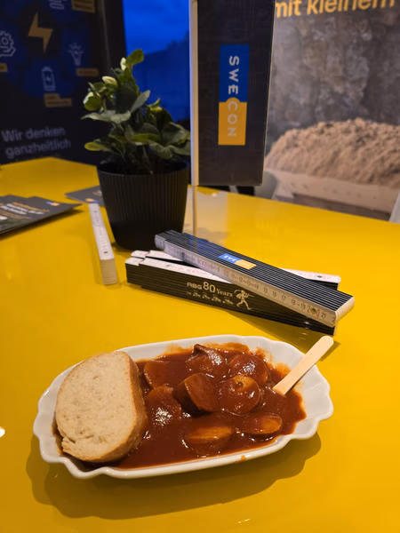 Currywurst mit Sauce und Brot auf einem gelben Tisch mit Swecon-Unterlagen