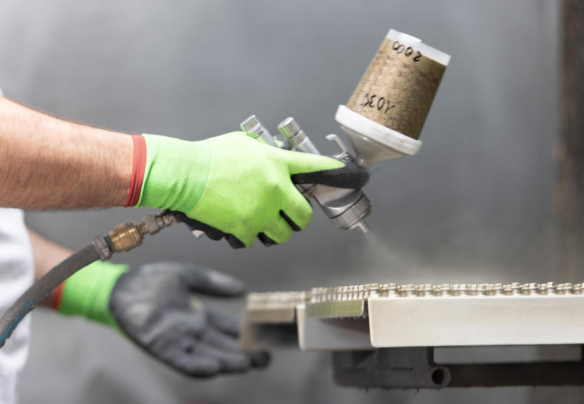 Hand mit Schutzhandschuh führt eine Lackierpistole über aufgereihte Befestiger.