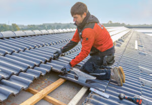 Monteur verschraubt eine Solar-Trägerpfanne auf einem mit grauen Dachpfannen eingedeckten Dach.