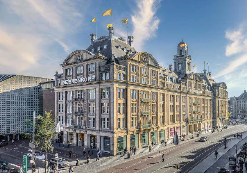 Gesamtansicht des Kaufhauses De Bijenkorf mit gegliederter Fassade, Dachaufbauten und gelben Fahnen.