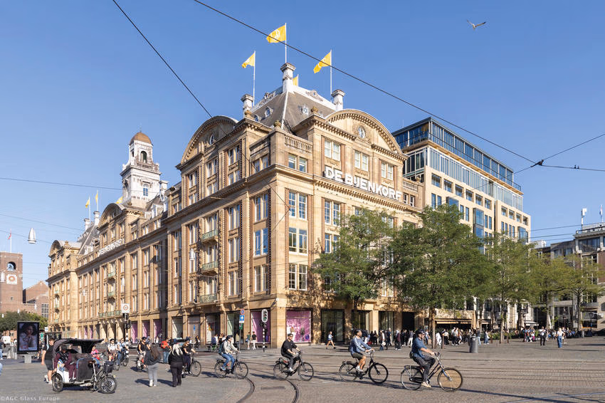 Kaufhaus De Bijenkorf mit gelben Fahnen, davor Radfahrer und Straßenbahnoberleitungen.