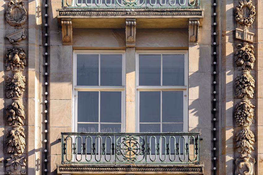 Zwei unterteilte Fenster hinter einem grünen Balkongeländer mit goldfarbenem Ornament.