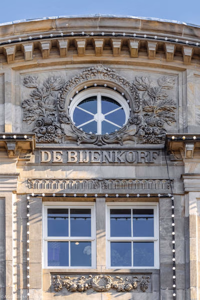 Rundes Fenster mit weißer Unterteilung über dem Schriftzug De Bijenkorf und zwei rechteckigen Fenstern.