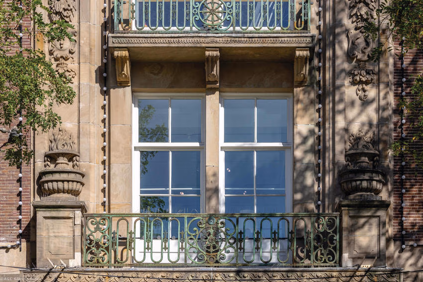 Weiße Fenster mit Spiegelungen von Bäumen hinter einem grünen, ornamentierten Balkongeländer.