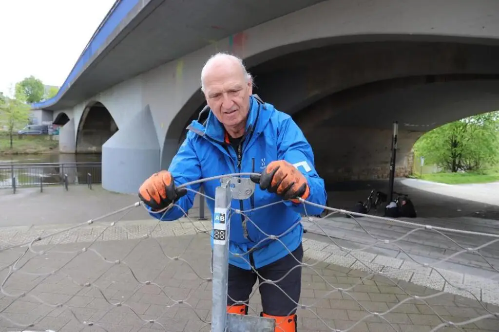 Aquaburg-Mitarbeiter Josef Winkels lässt das Stahlnetz in einen Pfosten einrasten. Als nächstes folgt die Plane. Im Hintergrund fließt ruhig die Ems, die an Weihnachten vergangenen Jahres an manchen Flussabschnitten jedoch bedrohlich über die Ufer trat.© Kerstin Heemann/DBU